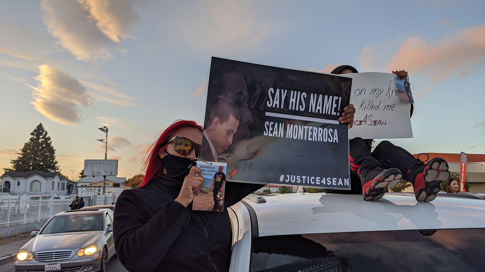 A young woman with red hair wearing sunglasses and a black face mask holds a sign that reads "Say his name! Sean Monterrosa."