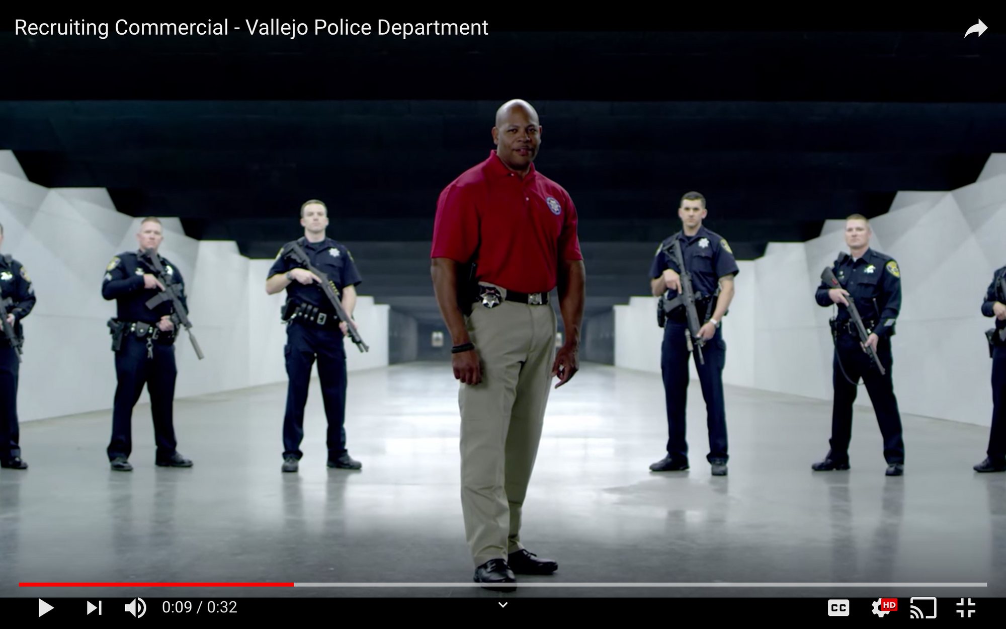 A police officer in a red polo shirt stands in the center of a room flanked by three officers in uniform on each side of him, all with guns in their hands.